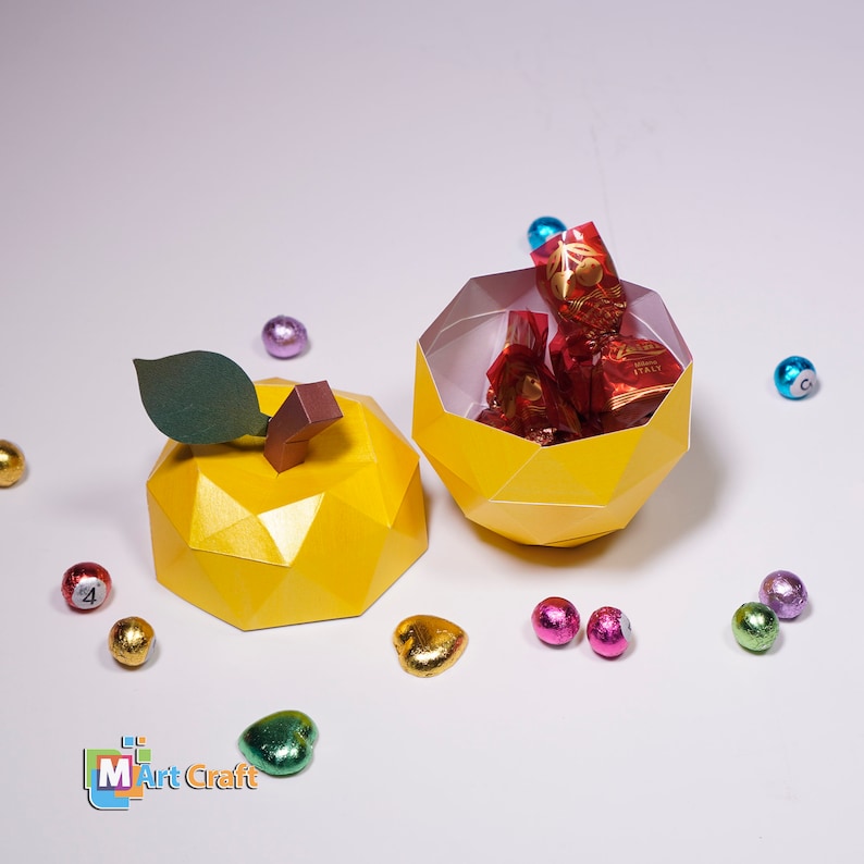 K&ouml;nnte beinhalten: Eine gelbe Papierbastel-Apfelbox mit einem gr&uuml;nen Blatt und einem braunen Stiel. Die Box ist ge&ouml;ffnet und mit rot und gold verpackten Pralinen gef&uuml;llt. Die Box ist von kleinen, bunten, in Folie verpackten Schokoladeneiern umgeben.