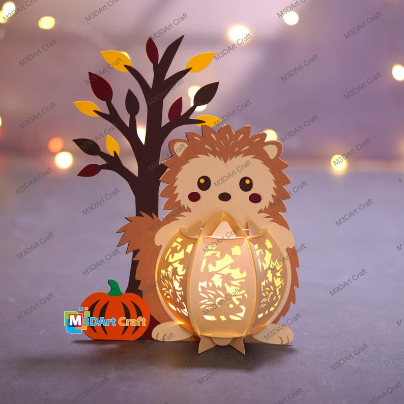 May include: A papercraft autumn scene featuring a hedgehog holding a glowing pumpkin lantern. A tree with fall-colored leaves stands behind the hedgehog, and a small pumpkin sits to the left. The text "M3DArt Craft" is visible.