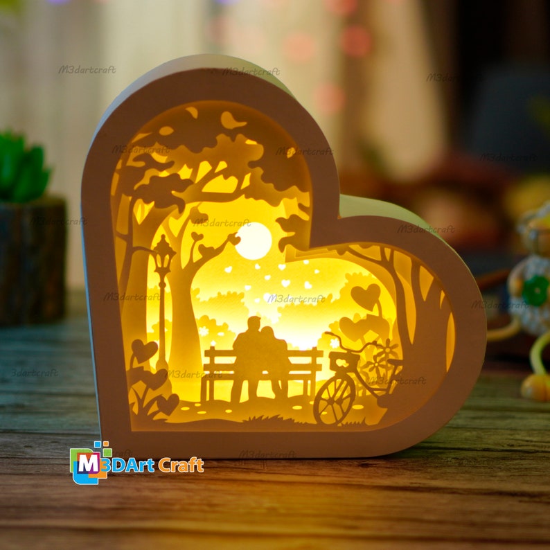May include: A white wooden heart-shaped lamp with a cutout design of a couple sitting on a bench in a park with a bicycle and a moon. The lamp is lit up with a warm yellow light.