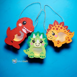Può includere: Tre lanterne di carta a forma di dinosauro appese, di colore rosso, verde e arancione. Il dinosauro rosso è a sinistra, il triceratopo verde al centro e lo stegosauro arancione a destra. Ognuna presenta una finestra ritagliata e uno spago.