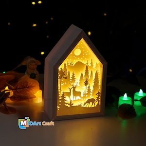 May include: A white wooden house-shaped night light with a cut-out design of a winter forest scene with deer and a fox. The light is illuminated from the inside, casting a warm glow.