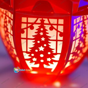 Puede incluir: Linterna roja de Navidad con un dise&ntilde;o recortado de un &aacute;rbol de Navidad y estrellas. La linterna est&aacute; iluminada desde adentro, creando un brillo c&aacute;lido.