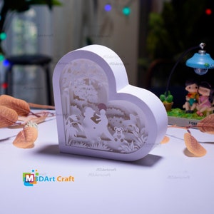 May include: White heart-shaped papercraft lamp with a cut-out design of a mother and child in a forest setting. The lamp is lit from behind, creating a warm glow. The text "M3DArt Craft" is visible at the bottom of the image.