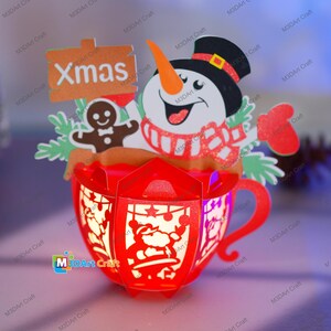 Puede incluir: Una taza de Navidad de papel rojo con un mu&ntilde;eco de nieve, un hombre de jengibre y un cartel que dice "Xmas". La taza est&aacute; decorada con purpurina y tiene una luz en el interior.