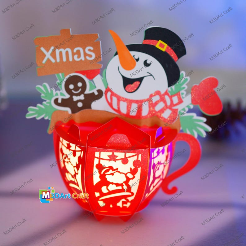 Puede incluir: Una taza de Navidad de papel rojo con un mu&ntilde;eco de nieve, un hombre de jengibre y un cartel que dice "Xmas". La taza est&aacute; decorada con purpurina y tiene una luz en el interior.