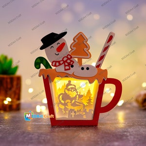 K&ouml;nnte beinhalten: Eine beleuchtete Weihnachts-Holztasse mit einem Schneemann, einem Zuckerstangen und einem Weihnachtsbaum oben drauf. Die Tasse hat eine beleuchtete Szene im Inneren mit dem Weihnachtsmann in einem Schlitten mit Rentieren.