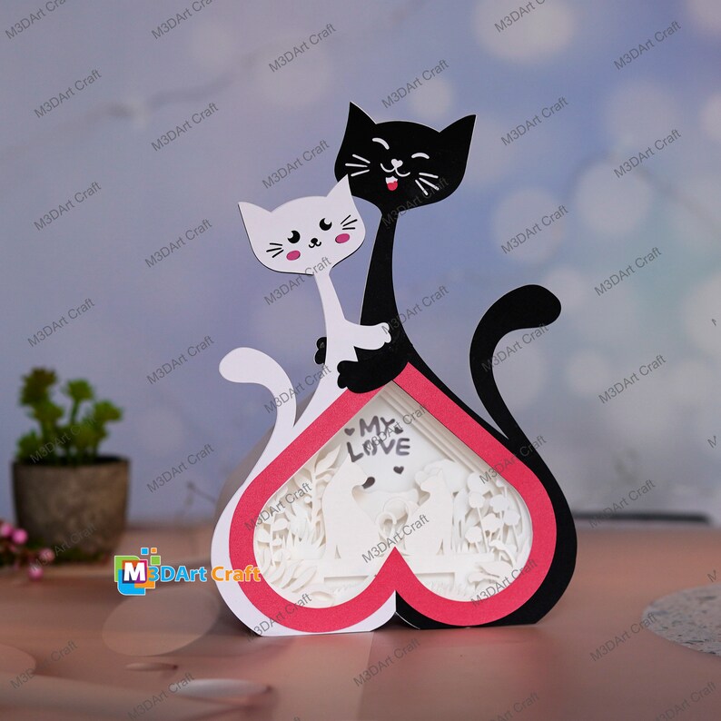 May include: A white and black papercraft of two cats in a heart-shaped frame. The cats are holding each other and the frame says "My Love".