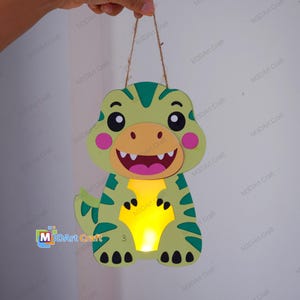 Op de afbeelding: Een lichtgroene dinosaurusvormige hanglamp met donkergroene strepen, een beige gezicht, roze wangen en een wijd open mond. Het midden van het dinosauruslichaam gloeit met een warm geel licht. Het item hangt aan een dun bruin touwtje.