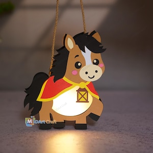 Peut inclure: Une décoration suspendue en bois en forme de cheval de dessin animé avec un corps marron, une crinière et une queue noires, et un visage blanc. Le cheval porte une cape rouge à bordure jaune et un motif de lanterne marron. Une lumière brille.