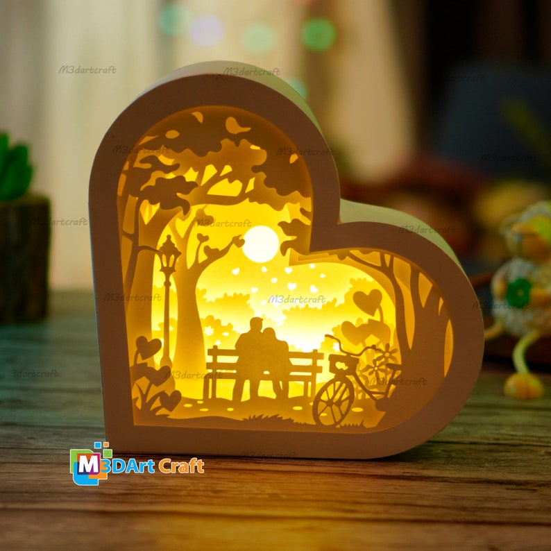 May include: A white wooden heart-shaped lamp with a cutout design of a couple sitting on a bench under a tree with a bicycle in the foreground. The lamp is illuminated from within, casting a warm glow.