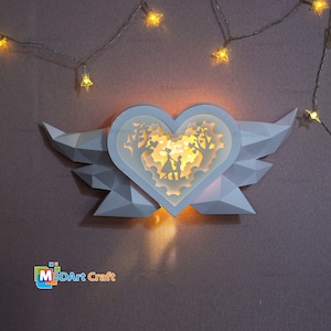 Puede incluir: Un corazón blanco de papel con alas, iluminado desde el interior. El corazón presenta un diseño recortado de una pareja de pie en un bosque con las palabras "Love You" encima.