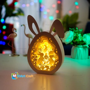 Peut inclure: Un œuf de Pâques en papier découpé blanc avec des oreilles de lapin, éclairé de l'intérieur. L'œuf est décoré d'un motif découpé de lapins, de fleurs et de feuilles. La lumière brille à travers les découpes, créant une lueur chaleureuse.