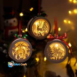 以下が含まれることがあります： 複雑な切り抜きデザインが施された紙製のクリスマスオーナメント3つ。オーナメントには、森の中の鹿、キリスト降誕の場面、雪だるまを持ったサンタクロースが描かれています。オーナメントは内側から照らされ、温かい光を放っています。