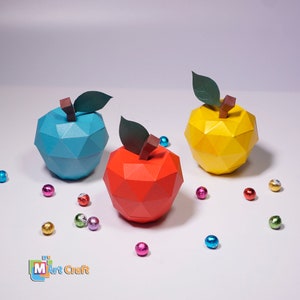 K&ouml;nnte beinhalten: Drei Origami-&Auml;pfel aus Papier in Blau, Gelb und Rot. Die &Auml;pfel liegen auf einer wei&szlig;en Oberfl&auml;che mit kleinen, bunten, gl&auml;nzenden Kugeln, die um sie herum verstreut sind. Der Text "M Art Craft" befindet sich in der linken unteren Ecke des Bildes.