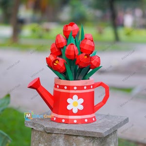 May include: A red paper watering can filled with red paper tulips and green leaves. The watering can has a white flower design and a pink band with white dots. The image is set on a stone surface.