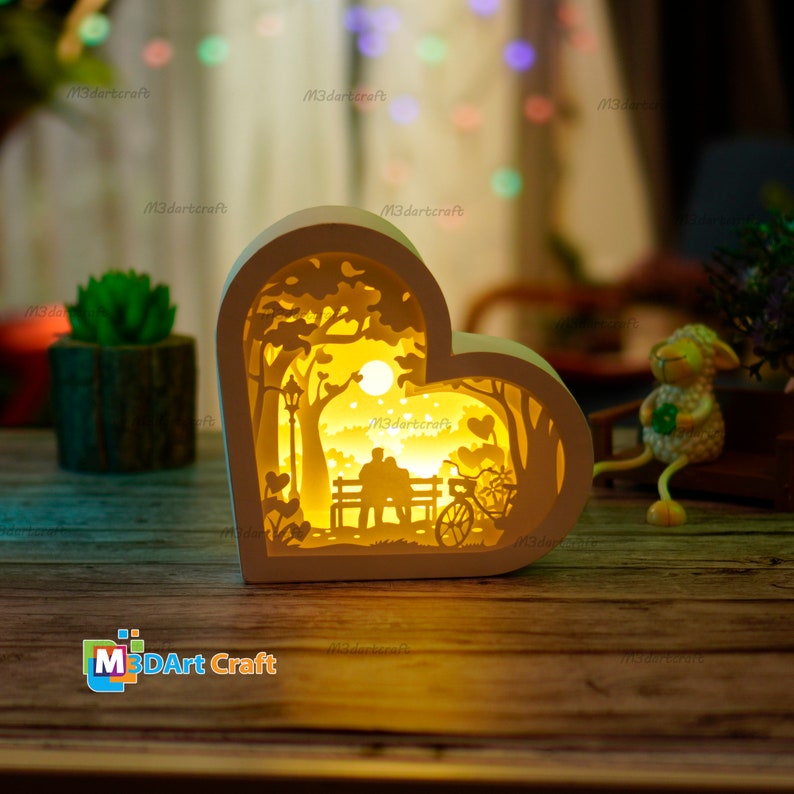 May include: A white wooden heart-shaped lamp with a cutout design of a couple sitting on a bench in a park with a bicycle and a tree. The lamp is lit up and casting a warm glow.