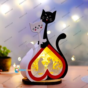 May include: A wooden heart-shaped lamp with a black and white cat silhouette on the front. The lamp has a red and white heart design and a cutout of two cats inside the heart. The word "love" is visible in the cutout.