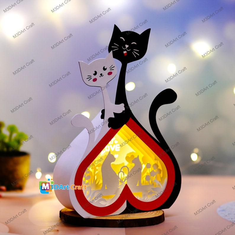 May include: A wooden heart-shaped lamp with a black and white cat silhouette on the front. The lamp has a red and white heart design and a cutout of two cats inside the heart. The word "love" is visible in the cutout.