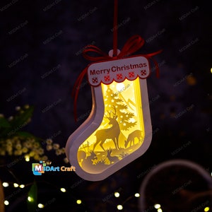 May include: White Christmas stocking ornament with a cut-out design of a deer, a tree, and other woodland creatures. The stocking is illuminated from within and has a red ribbon with the words "Merry Christmas" on it.