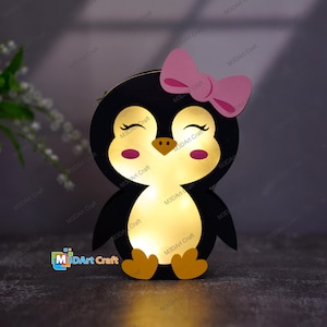 Może przedstawiać: Drewniana lampka nocna w kształcie pingwina z różową kokardką, czarnymi oczami i jasnożółtym blaskiem. Pingwin siedzi na czarnej powierzchni.