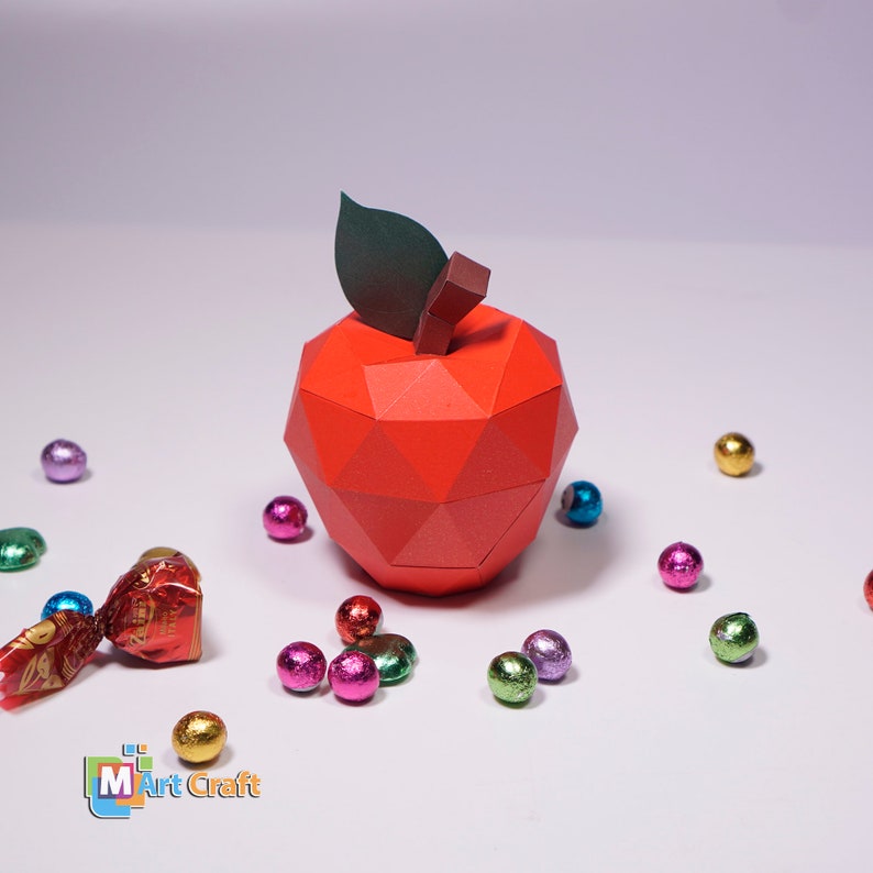 K&ouml;nnte beinhalten: Ein roter Papierapfel mit einem gr&uuml;nen Blatt, umgeben von bunten, in Folie verpackten S&uuml;&szlig;igkeiten. Der Apfel ist eine geometrische Form mit vielen Facetten. Die S&uuml;&szlig;igkeiten sind auf einer wei&szlig;en Oberfl&auml;che verstreut.