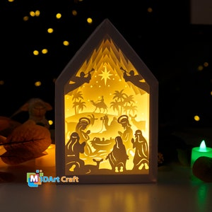 May include: A white wooden nativity scene with a light inside, depicting the birth of Jesus with Mary, Joseph, and the baby Jesus in a manger. The scene is illuminated from behind, creating a warm glow. There are palm trees and a star in the background.