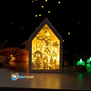 May include: A white wooden house-shaped light-up decoration with a cut-out design of a winter village scene, including houses, trees, a snowman, and Santa Claus in a sleigh pulled by reindeer. The light shines through the cut-outs, creating a warm glow.