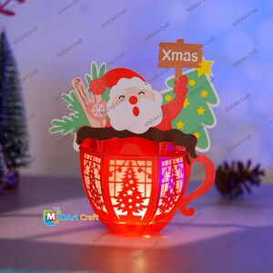 Puede incluir: Una linterna de papel roja con forma de taza con un dise&ntilde;o de Pap&aacute; Noel y un &aacute;rbol de Navidad. La linterna tiene una luz en el interior y dice "Xmas" en un cartel.