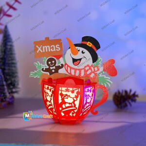 Puede incluir: Una taza de Navidad de papel roja, iluminada, con un mu&ntilde;eco de nieve, un hombre de jengibre y un cartel que dice "Xmas".