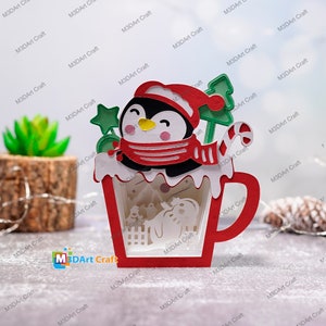 K&ouml;nnte beinhalten: Eine rote und wei&szlig;e 3D-Papiercraft-Tasse mit einem Pinguin, der einen roten Schal und eine Weihnachtsm&uuml;tze tr&auml;gt. Die Tasse ist mit einem gr&uuml;nen Stechpalmenzweig und einem Zuckerstangen verziert.