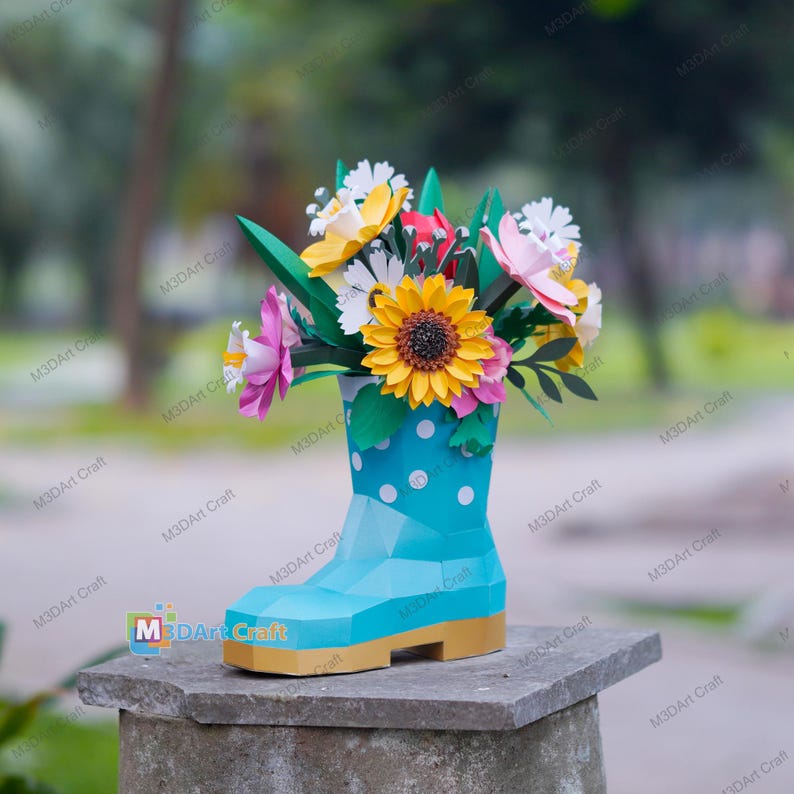 May include: A papercraft floral arrangement featuring a blue boot vase with white polka dots, holding colorful paper flowers. The flowers include a large yellow sunflower, pink and white blossoms, and green leaves. The boot has a tan sole.
