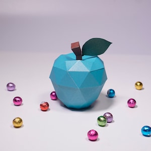 K&ouml;nnte beinhalten: Ein blauer Origami-Apfel mit einem gr&uuml;nen Blatt und einem braunen Stiel. Der Apfel ist von bunten, in Folie verpackten Schokoladeneiern umgeben.