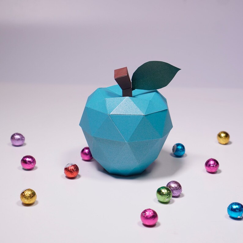 K&ouml;nnte beinhalten: Ein blauer Origami-Apfel mit einem gr&uuml;nen Blatt und einem braunen Stiel. Der Apfel ist von bunten, in Folie verpackten Schokoladeneiern umgeben.