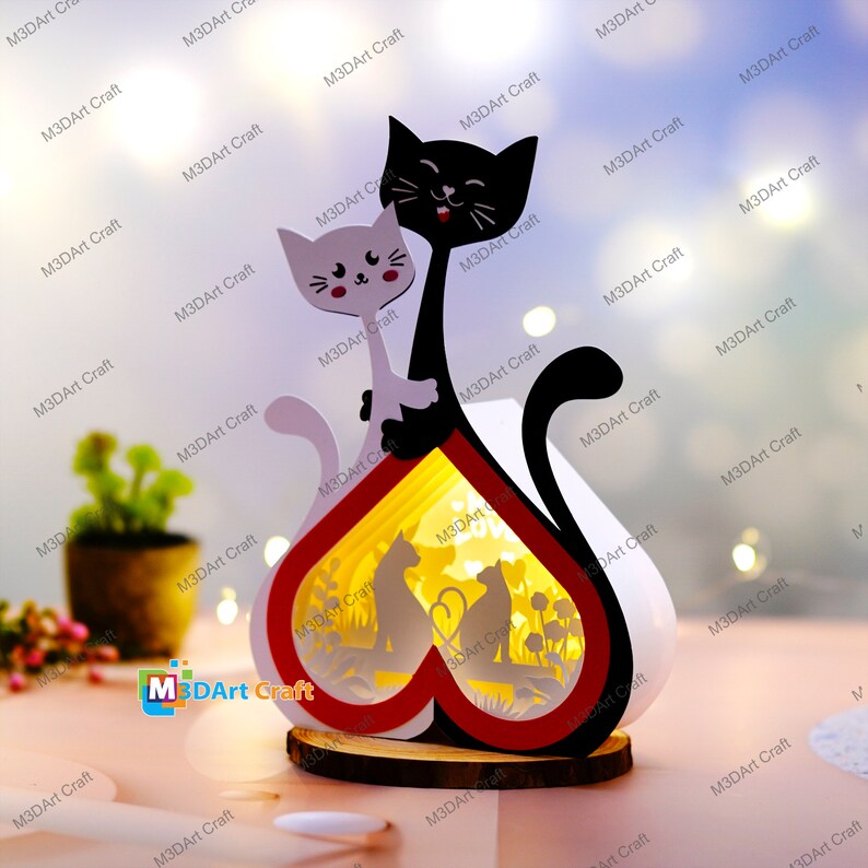 May include: A wooden heart-shaped lamp with a black and white cat silhouette on each side. The lamp has a red and white heart design and a light inside that illuminates a scene of two cats sitting in a field of flowers.
