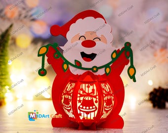 Op de afbeelding: Een rode en witte kerstman-uitsparing die een lichtsnoer vasthoudt. De uitsparing is een lantaarn met een licht dat door het ontwerp schijnt.