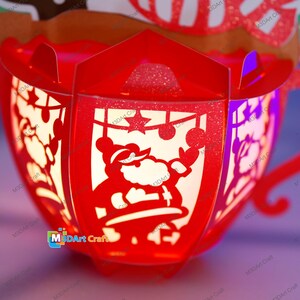 Puede incluir: Una linterna de papel roja con un dise&ntilde;o recortado de Pap&aacute; Noel, una estrella y adornos. La linterna est&aacute; iluminada desde el interior, creando un brillo c&aacute;lido.