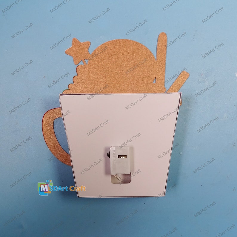 K&ouml;nnte beinhalten: Ein wei&szlig;er Lichtschalter mit einer goldfarbenen dekorativen Lichtschalterabdeckung, die ein Cartoon-Bild einer Tasse Kaffee mit einem Stern obenauf zeigt. Die Schalterabdeckung ist mit "M3DArt Craft" beschriftet.
