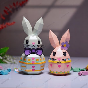 Peut inclure: Deux lapins de Pâques en papier. L'un est blanc avec un nœud papillon violet, l'autre est rose avec une fleur. Les deux ont des motifs géométriques et sont décorés de cœurs et de fleurs. Les lapins sont sur une surface claire.