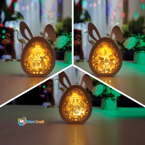 Może przedstawiać: Biała drewniana lampka w kształcie jajka wielkanocnego z uszami króliczka. Jajko ma wycięty wzór króliczka, motyla i jajek. Lampka jest oświetlona ciepłym żółtym światłem. Wewnątrz jajka widoczny jest napis "Happy Easter".