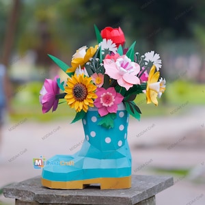 May include: A colorful paper flower arrangement in a turquoise boot-shaped vase with white polka dots. The bouquet includes a yellow sunflower, pink and white flowers, and green leaves. The vase has a yellow base.