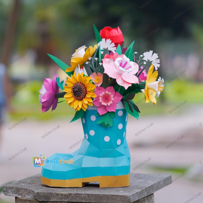 May include: A colorful paper flower arrangement in a turquoise boot-shaped vase with white polka dots. The bouquet includes a yellow sunflower, pink and white flowers, and green leaves. The vase has a yellow base.