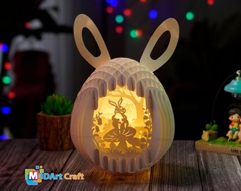 May include: A white papercraft Easter egg with bunny ears, illuminated from within, depicting a rabbit sitting on a mushroom in a forest scene. The egg is made of layered paper and has a 3D effect.