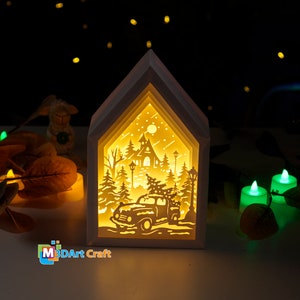 May include: A white wooden house-shaped lamp with a 3D paper cut-out design of a winter scene. The scene includes a church, trees, a car with a Christmas tree on top, and a moon in the sky. The lamp is lit up, creating a warm glow.