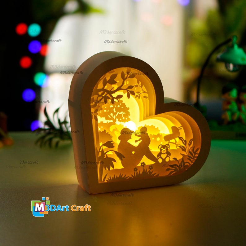 May include: A white heart-shaped paper lamp with a 3D layered design. The lamp features a silhouette of a mother and child sitting in a garden with a tree, flowers, and a teddy bear. The lamp is illuminated from within, casting a warm glow.