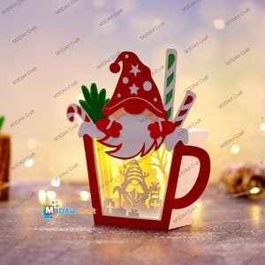 K&ouml;nnte beinhalten: Eine rote und wei&szlig;e Holzfigur eines Weihnachtszwergs, die einen Zuckerstock in einer roten und wei&szlig;en Tasse mit einem Licht darin h&auml;lt. Die Tasse ist mit einem gr&uuml;nen Zweig von Stechpalme und einem roten und wei&szlig;en Zuckerstock verziert.