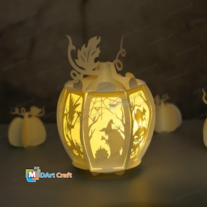 May include: A white papercraft pumpkin lantern with a light inside. The lantern is decorated with a witch, a spider web, and a tree. The lantern is lit up and the light shines through the cutouts in the paper.