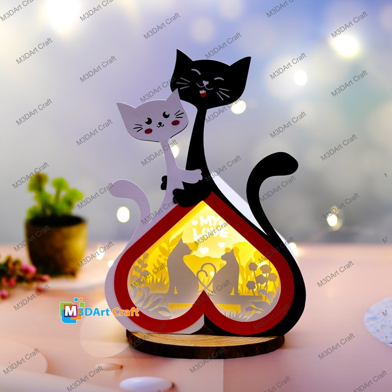 May include: A wooden heart-shaped lamp with a red and white border. The lamp features a cutout design of two cats sitting on a bench with a heart and the words "My Love" inside the heart. The lamp is lit from behind, creating a warm glow.