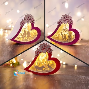 Puede incluir: Tres cajas de madera en forma de corazón iluminadas con intrincados diseños recortados. Las cajas presentan escenas de pájaros, parejas y una familia con mascotas. Las cajas están iluminadas desde el interior, creando un brillo cálido.