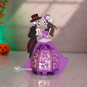 Peut inclure: Une lanterne en papier représentant un couple de squelettes en tenue de soirée, la femme portant une robe violette et l'homme un costume noir et un chapeau haut de forme. La lanterne a un motif floral et est éclairée de l'intérieur.