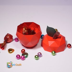 K&ouml;nnte beinhalten: Eine rote Papier-Apfelbox mit einem gr&uuml;nen Blatt oben. Die Box ist ge&ouml;ffnet und mit verpackten Pralinen gef&uuml;llt. Es gibt mehrere kleine, bunte, in Folie verpackte S&uuml;&szlig;igkeiten, die um die Box herum verstreut sind.